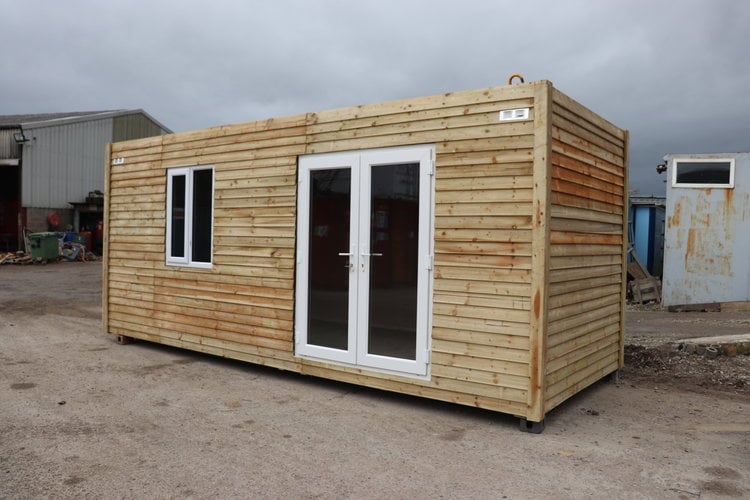 Wooden mobile cabin with white-framed windows and dark double doors on a gravel industrial yard